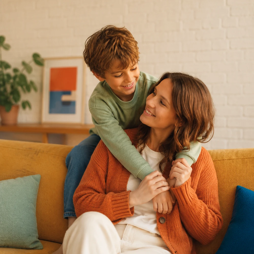 A young boy hugs his smiling mother from behind on a mustard-yellow couch, both sharing a warm, affectionate moment in a cozy living room.